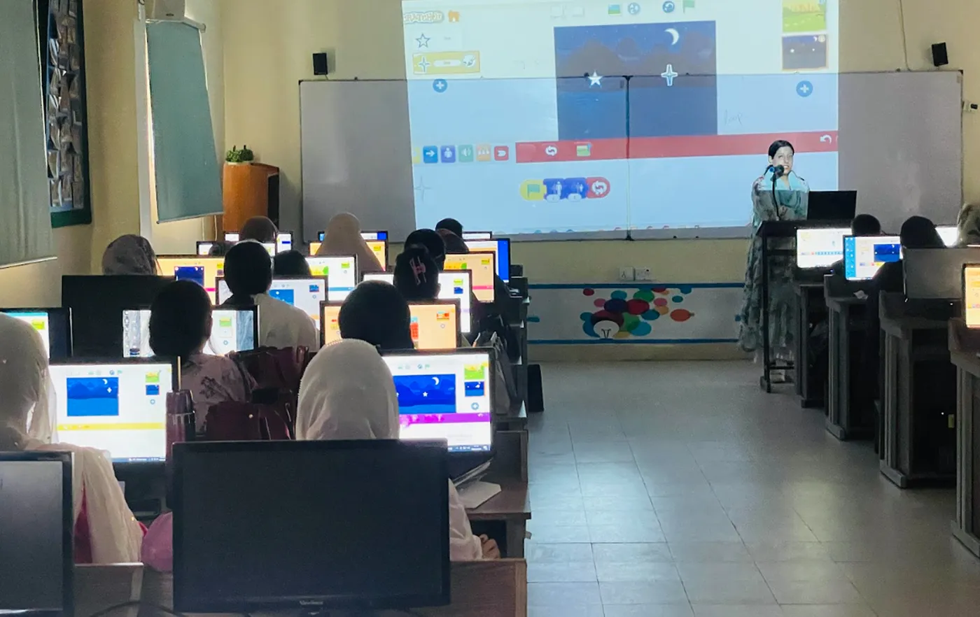 A Scratch lesson projected in a computer lab full of learners